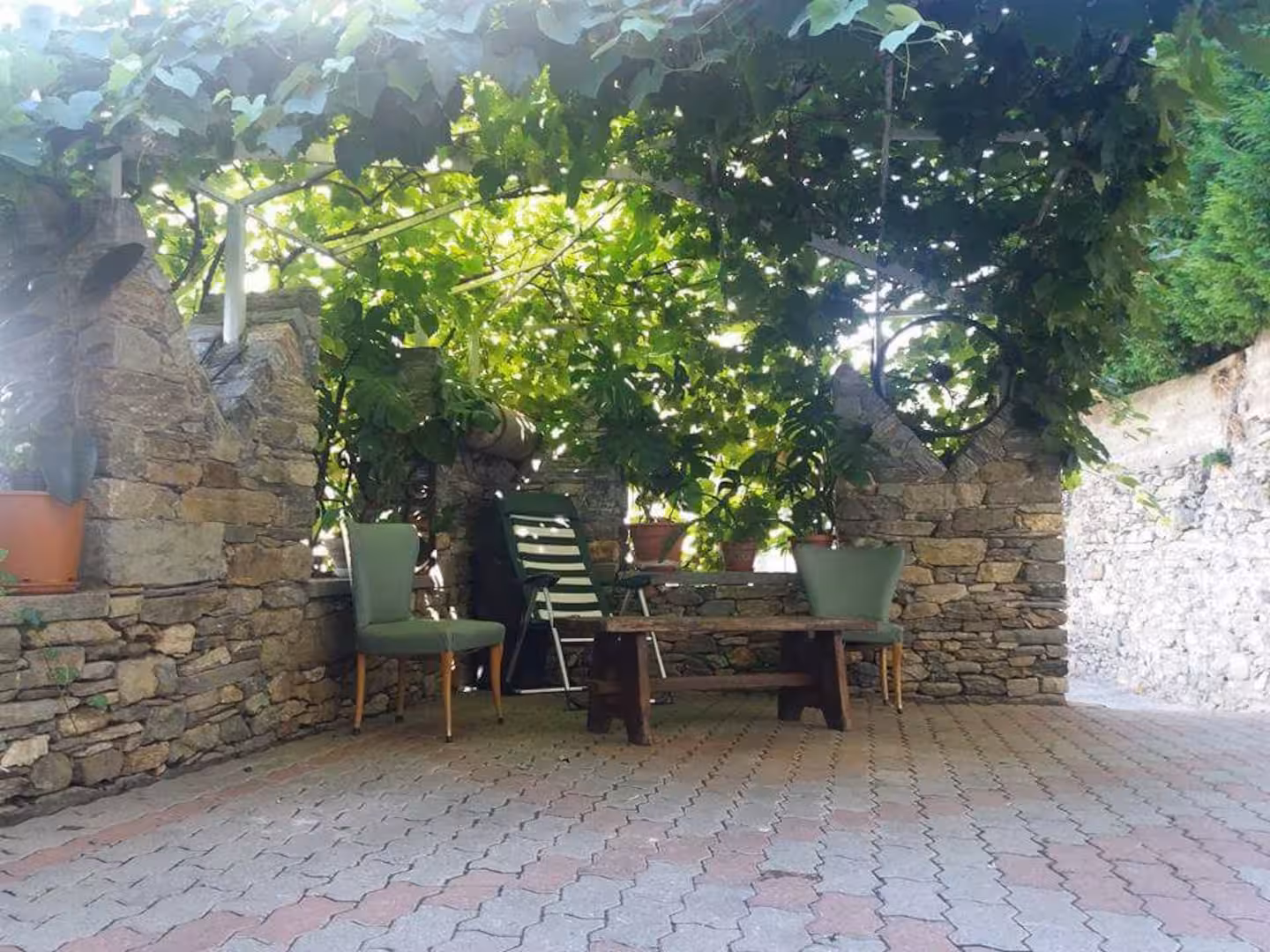 Outdoor terrace under vine pergola with stone wall and wooden table and chairs
